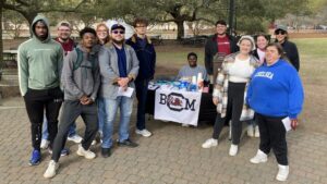 A large group of students pose on either side of a table with a BCM table cloth on the quad of the University of South Carolina